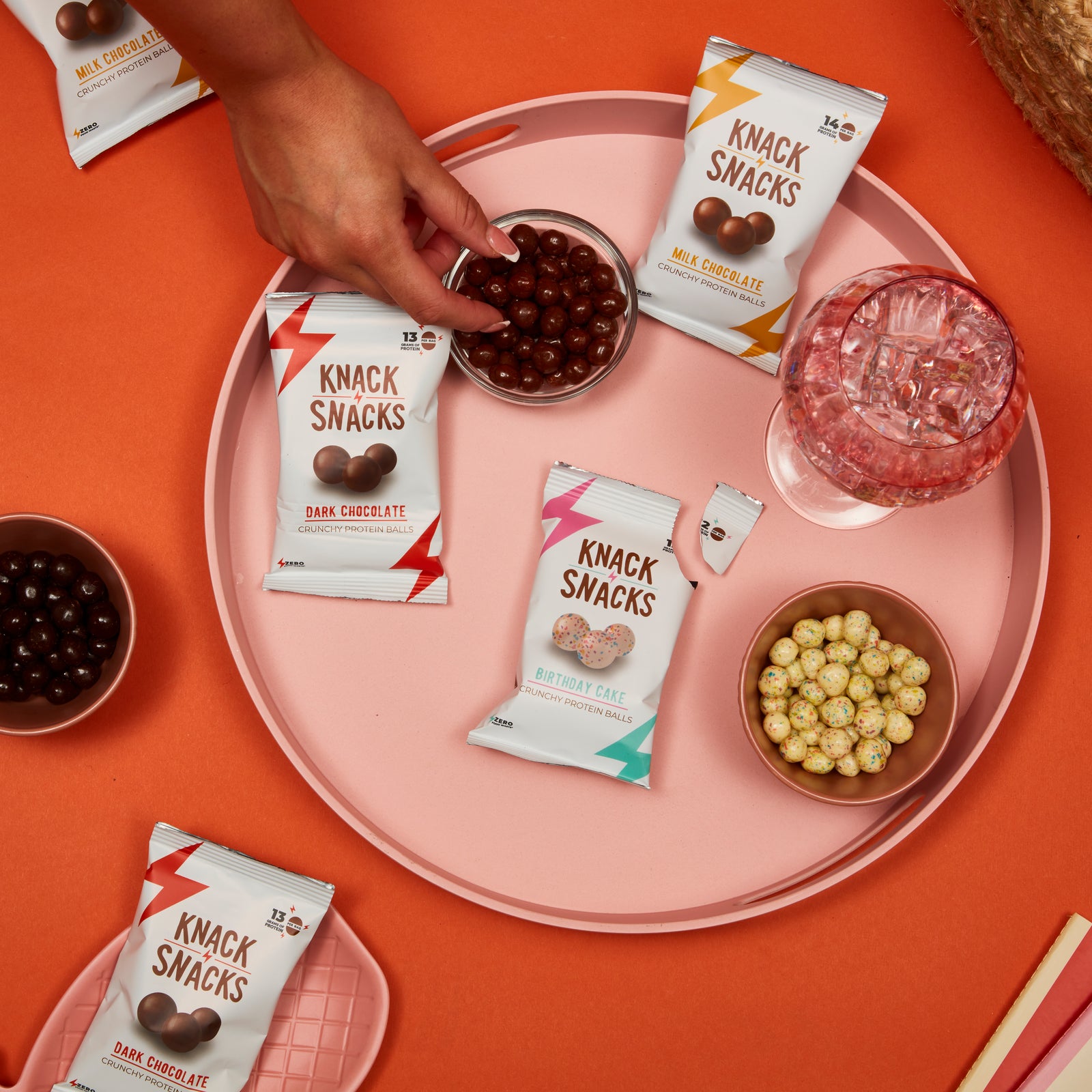 Packs and bowls of Knack Snacks crunchy protein balls in milk chocolate, dark chocolate and salted caramel, styled on a pink plate